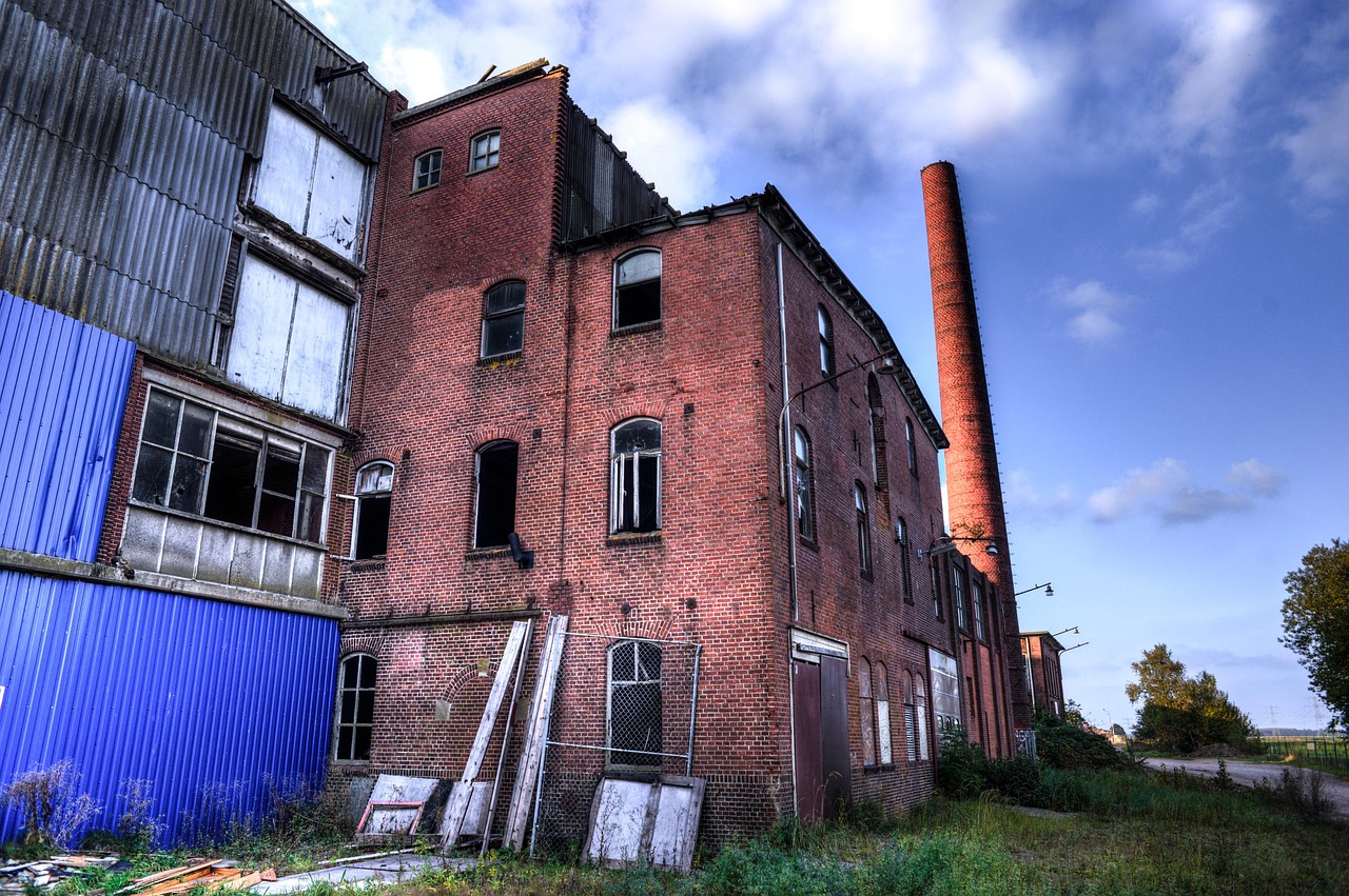 Ancienne usine en briques abandonnée, avec sa haute cheminée et un terrain envahi d'herbes folles