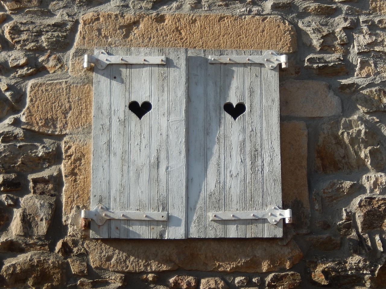volets de bois blanchis fermés, percés de deux cœurs, sur un mur de maison en pierre